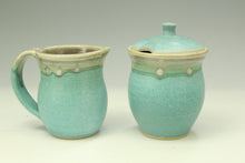 Load image into Gallery viewer, A ceramic pitcher and matching lidded container with a glossy aqua glaze and beige rim, photographed on a light gray studio background.