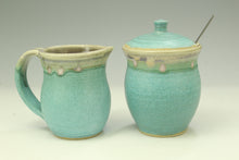 Load image into Gallery viewer, A ceramic pitcher and matching lidded container with a spoon, both glazed in a light turquoise/aqua color with a rustic beige and green band near the rim, photographed on a neutral light gray background.